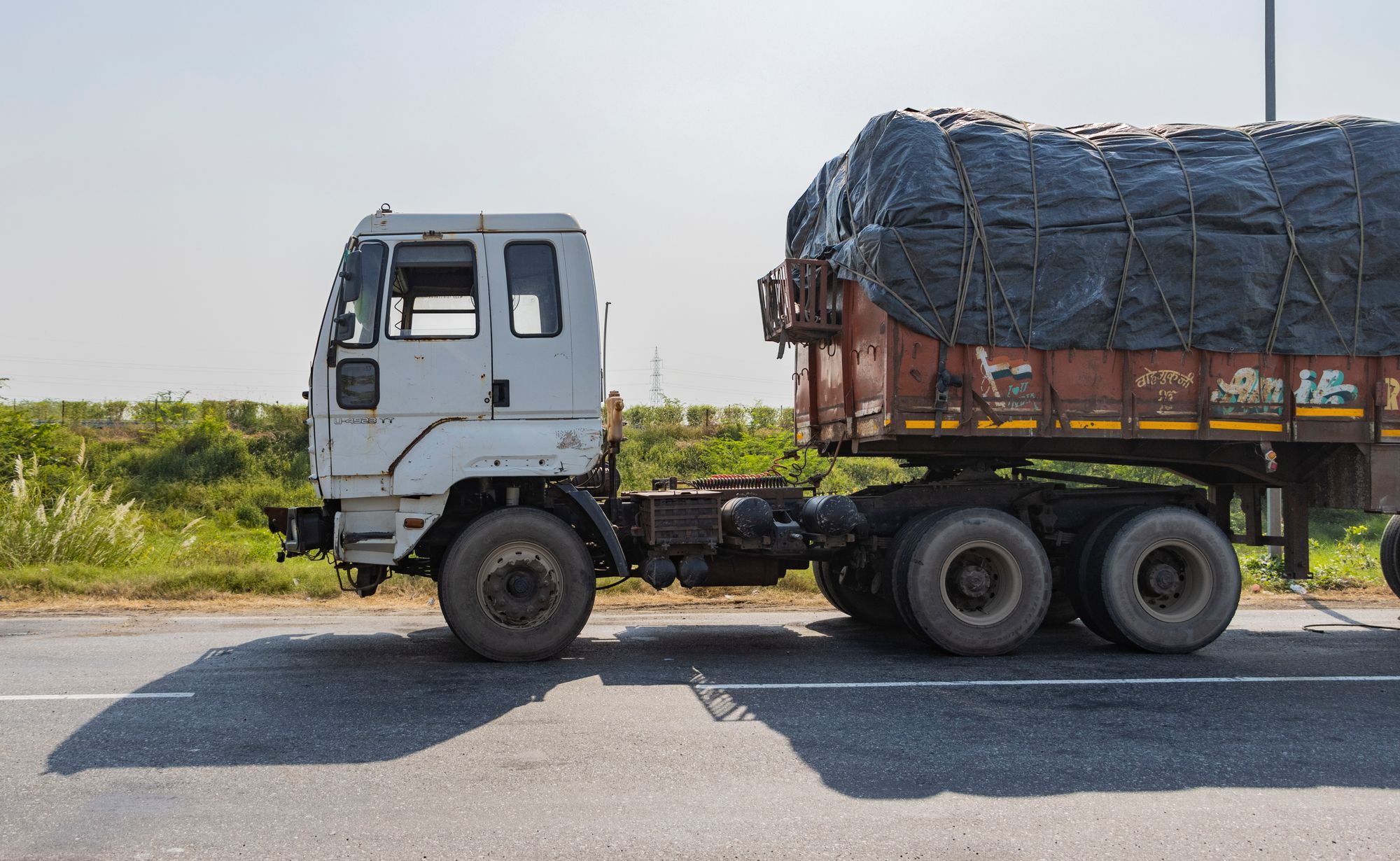 A goods truck with a tarpaulin-covered trailer seen from the side, standing on an Indian highway.